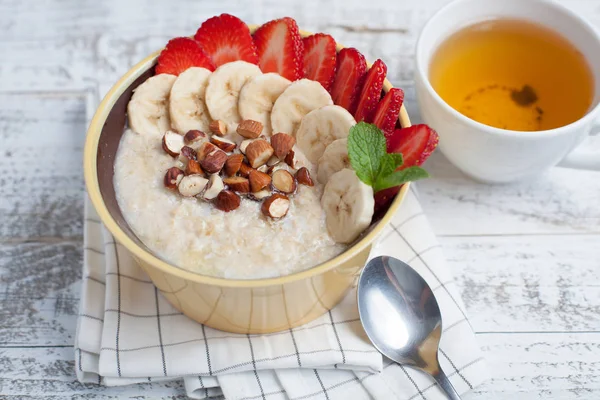 Bowl of oatmeal with a banana, strawberries, almonds, hazelnuts and butter on a rustic table. Hot and a healthy dish for Breakfast, top view — Stock Photo, Image