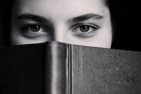 A girl with big eyes hides behind a book. Close-up portrait. Black and white photo — Stock Photo, Image