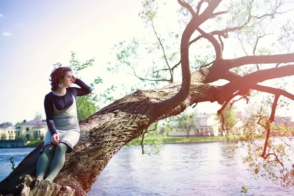 Uma jovem se senta em um galho de árvore perto da água e toca seu cabelo. Retrato de primavera ao pôr-do-sol — Fotografia de Stock
