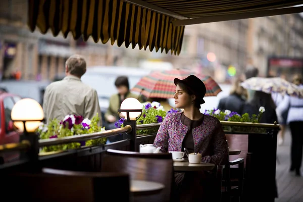 Schöne junge Frau mit Hut und einer Tasse Tee, die in einem Café sitzt und Passanten ansieht — Stockfoto