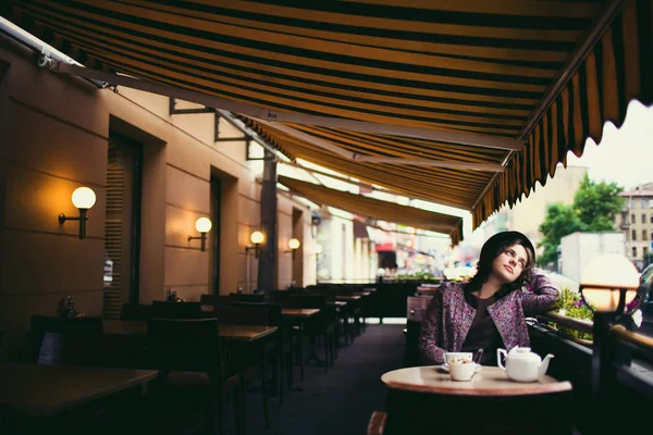 Mulher bonita em um chapéu com uma xícara de chá sentado em um café, olhando para as pessoas que passam por — Fotografia de Stock