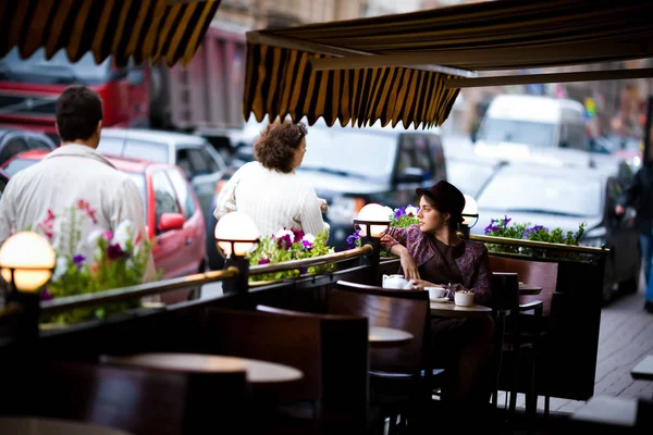 Belle jeune femme dans un chapeau avec une tasse de thé assis dans un café, regardant les gens qui passent — Photo