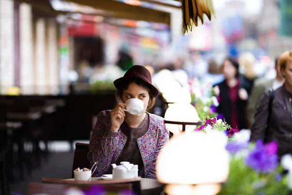 Eine schöne junge Frau mit Hut sitzt auf einer Terrasse in einem Café, trinkt Tee und beobachtet Passanten — Stockfoto