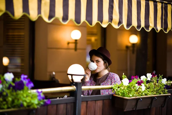 Schöne junge Frau mit Hut sitzt auf einer Terrasse in einem Café, trinkt Tee und blickt in die Kamera — Stockfoto