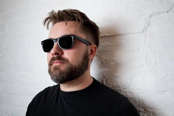 Retrato de un hombre serio barbudo con gafas de sol y ropa casual contra una pared de ladrillo blanco . — Foto de Stock