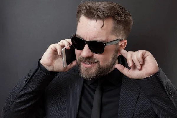 Un hombre de negocios con traje negro y gafas está hablando por teléfono. Él no oye a su interlocutor, por lo que se tapa la oreja libre con el dedo. Problema con la comunicación celular — Foto de Stock