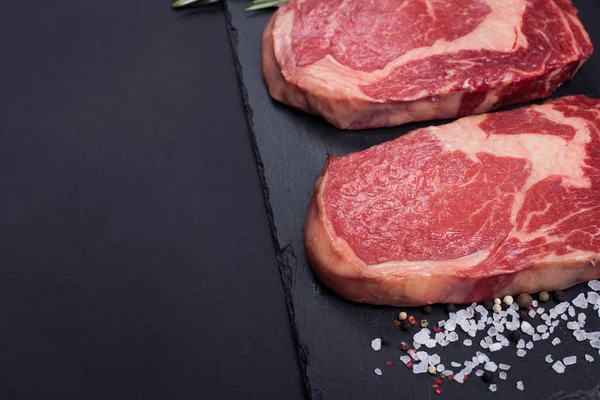 Dos carnes frescas de mármol crudo, filete negro Angus ribeye con especias sobre un fondo de piedra oscura . — Foto de Stock