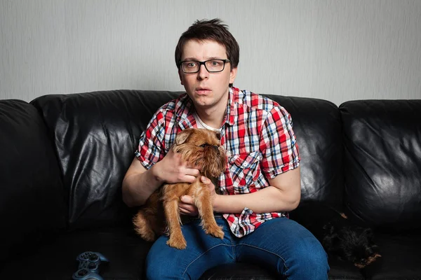 Closeup portrait, young man in the red shirt, sitting on black leather couch with two dogs, watching TV, holding remote, surprised at what he sees. He grabbed a red dog in surprise