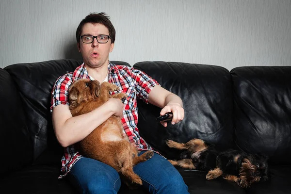 Retrato de cerca, joven con la camisa roja, sentado en un sofá de cuero negro con dos perros, viendo la televisión, sosteniendo el control remoto, sorprendido de lo que ve. Agarró un perro rojo sorprendido. — Foto de Stock