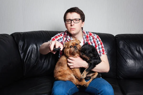 Портрет крупным планом, молодой человек в красной рубашке, сидит на черном кожаном диване с двумя собаками, смотрит телевизор, держит пульт дистанционного управления, удивляется тому, что он видит. Он взял собак на руки с удивлением — стоковое фото