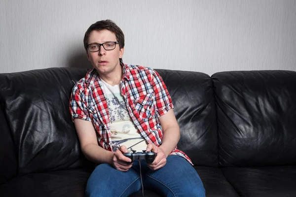 Молодой парень в очках и красной рубашке играет в видеоигры на джойстике, сидит на черном кожаном диване — стоковое фото