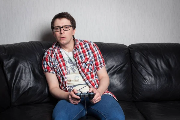 Jonge man met bril en rood shirt spelen van videogames op de joystick, zittend op een zwarte leerbank — Stockfoto