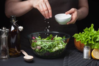 Woman chef in the kitchen preparing vegetable salad. Healthy Eating. Diet Concept. A Healthy Way Of Life. To Cook At Home. For Cooking. The girl sprinkles sea salt in a salad on a dark background clipart