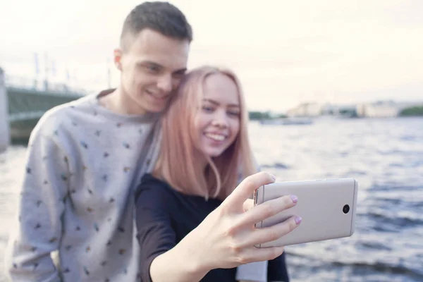 年轻夫妇拍摄自拍照与智能手机背景下的城市景观。在圣彼得堡的河堤 — 图库照片