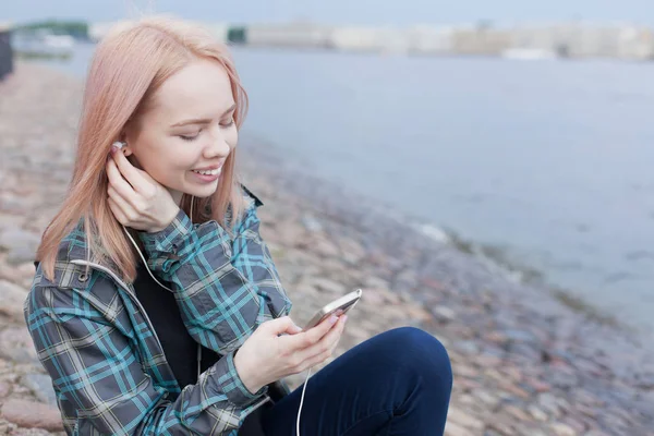 Joven chica hermosa sentada en la playa y escuchando música en su teléfono inteligente. Ella sonríe disfrutando de la música —  Fotos de Stock