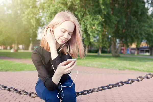 Jovem menina bonita senta-se e ouve música em seu smartphone. Ela sorri, aproveitando a música e endireita o cabelo — Fotografia de Stock