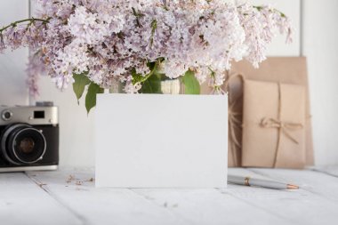 Blank white greeting card with lilac flowers bouquet and envelope with vintage camera on white wooden background. mock up clipart