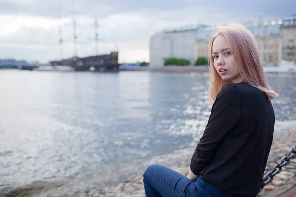 Menina bonita jovem senta-se à beira-mar e olhando para a câmera. No fundo está desfocado velho navio — Fotografia de Stock