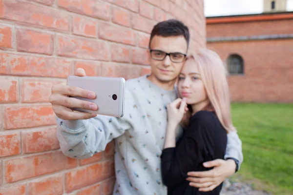 Genç bir çift bir selfie kırmızı tuğla duvar arka plan üzerinde smartphone ile fotoğraf çekmek. Mavi gözleri ve Arap görünüm kahverengi gözlü genç bir adam sarışın kız. — Stok fotoğraf