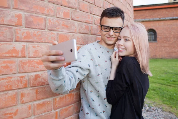 年轻夫妇拍摄自拍照与背景的红砖墙上的智能手机。蓝色的眼睛和一个年轻人的阿拉伯外观棕色的眼睛的金发女郎. — 图库照片