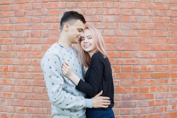 Счастливая молодая пара обнимается и улыбается на фоне красной кирпичной стены. Блондинка с голубыми глазами и молодой человек арабской внешности с карими глазами — стоковое фото