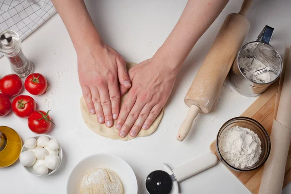 Manos masculinas preparando la masa para la pizza en primer plano de la mesa. En la mesa blanca están los tomates, las bolas de mozzarella, el aceite de oliva y la harina — Foto de Stock