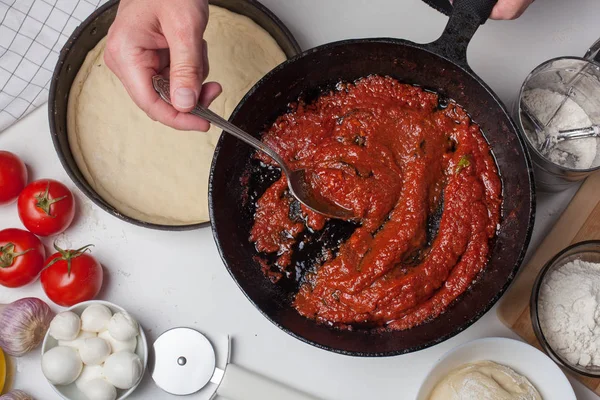 Chef macho sosteniendo una sartén con salsa de tomate para la pizza. En la mesa blanca están los tomates, las bolas de mozzarella, el aceite de oliva y la harina — Foto de Stock