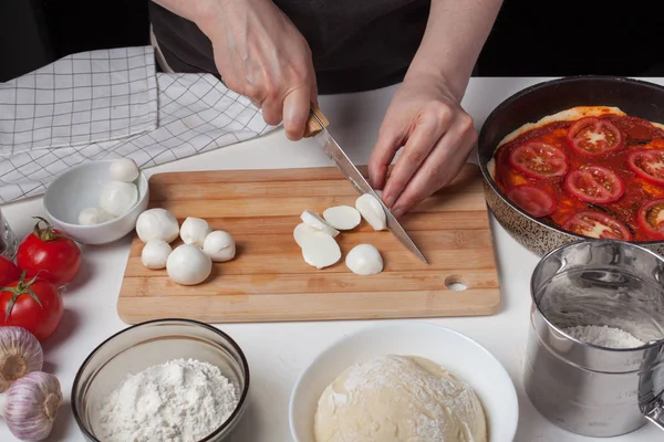 Una chef rebanada queso mozzarella para hacer una pizza italiana casera. En la mesa blanca hay mozzarella, masa de pizza y ajo. — Foto de Stock