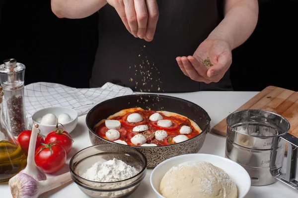 Las manos de la mujer cocinera espolvorear italiana pizza cruda margarita oreano sobre un fondo oscuro. En la mesa blanca se encuentran los ingredientes de mozzarella, ajo, masa de levadura, harina y aceite de oliva — Foto de Stock