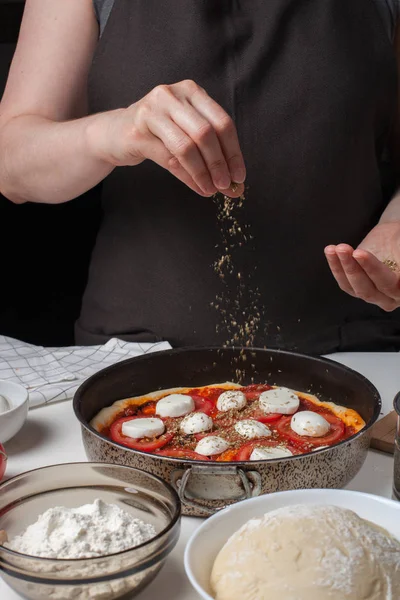 Las manos de la mujer cocinera espolvorear italiana pizza cruda margarita oreano sobre un fondo oscuro. En la mesa blanca se encuentran los ingredientes de mozzarella, ajo, masa de levadura, harina y aceite de oliva — Foto de Stock