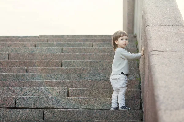 Un niño de ojos azules con el pelo largo parado frente a las escaleras de piedra. El concepto de crecer — Foto de Stock