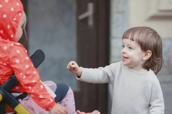 Счастливый ребенок с длинными светлыми волосами играет с маленькой девочкой сидящей в детской коляске — стоковое фото