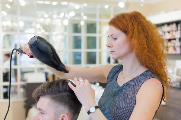 Un cliente varón cortándose el pelo. Chica peluquera seca mi cabello un chico joven y atractivo en un salón de belleza —  Fotos de Stock