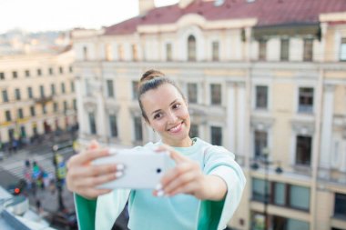Kahverengi saçlı sevimli seksi kız eski şehir Evin çatısı üzerinde duruyor ve smartphone bir selfie yapar. Şık kadın kameraya gülümseyen