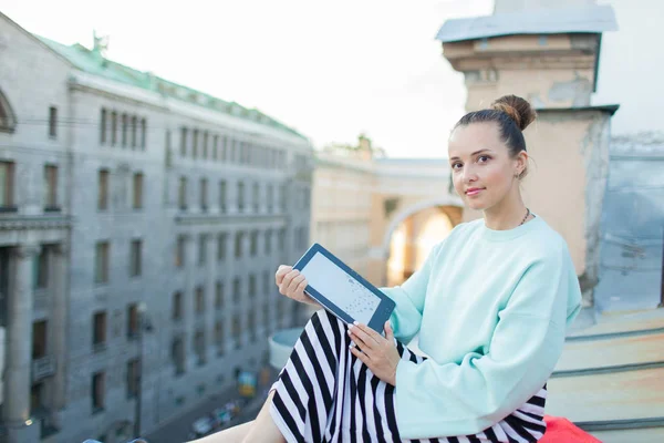 Menina atraente e elegante senta-se no telhado da casa na cidade velha e lê o e-book — Fotografia de Stock