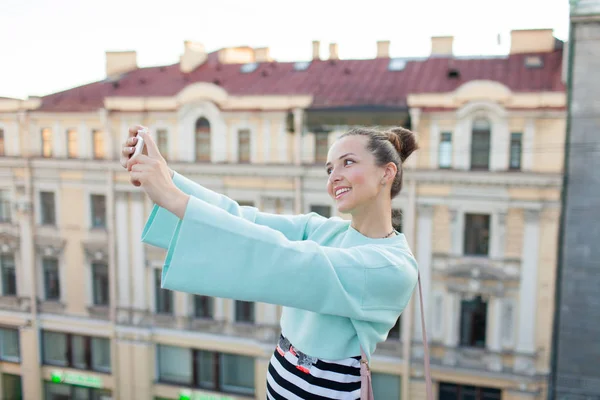 Menina sexy bonito com cabelos castanhos fica no telhado da casa na cidade velha e faz uma selfie em seu smartphone — Fotografia de Stock