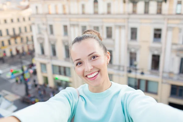 Menina sexy bonito com cabelos castanhos fica no telhado da casa na cidade velha e faz uma selfie em seu smartphone. Mulher elegante sorrindo para a câmera — Fotografia de Stock