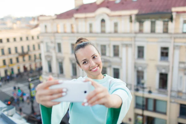 Kahverengi saçlı sevimli seksi kız eski şehir Evin çatısı üzerinde duruyor ve smartphone bir selfie yapar. Şık kadın kameraya gülümseyen — Stok fotoğraf