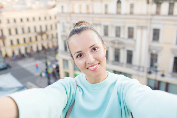 Menina sexy bonito com cabelos castanhos fica no telhado da casa na cidade velha e faz uma selfie em seu smartphone. Mulher elegante sorrindo para a câmera — Fotografia de Stock