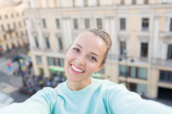 Menina sexy bonito com cabelos castanhos fica no telhado da casa na cidade velha e faz uma selfie em seu smartphone. Mulher elegante sorrindo para a câmera — Fotografia de Stock