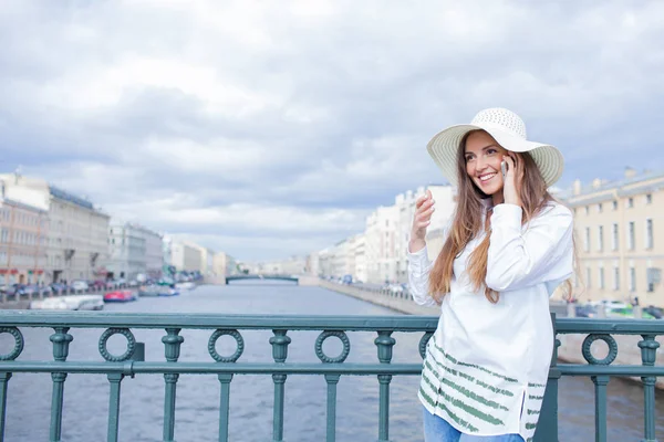 Uma menina bonita e sorridente em um chapéu branco com aba larga está de pé na ponte e falando ao telefone contra o fundo de nuvens azuis — Fotografia de Stock