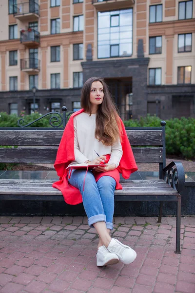 Atrakcyjna dziewczyna z długimi włosami siedzi na ławce, pokryte czerwoną kocem, w nowej dzielnicy mieszkaniowej i zapisuje jej myśli w czerwony Notes — Zdjęcie stockowe
