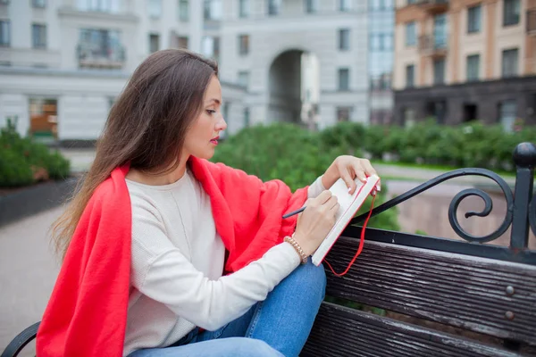 Chica atractiva con el pelo largo se sienta en un banco, cubierto con una manta roja, en un nuevo barrio residencial y escribe sus pensamientos en un cuaderno rojo —  Fotos de Stock