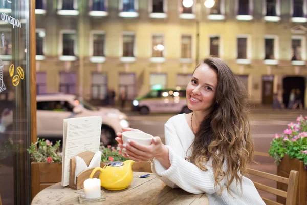 Menina atraente jovem sentado à noite em um café com uma xícara de chá para o pano de fundo de carros que passam e vida na cidade. Ela olha para a câmera e sorri — Fotografia de Stock