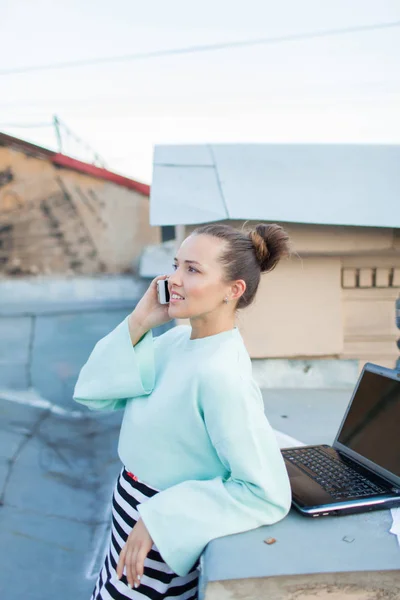 Bonita mulher de negócios falando ao telefone em pé no telhado da casa na cidade velha. Ao lado dele estão um laptop, smartphone e documentos em papel — Fotografia de Stock