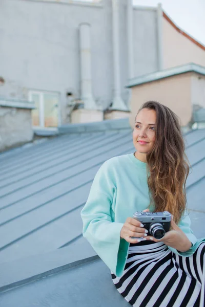 Look de moda, modelo jovem muito legal com câmera de filme retro. cabelo encaracolado ao ar livre. Fotógrafo menina elegante leva a cidade velha do telhado ao pôr do sol . — Fotografia de Stock