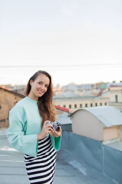 Mode-Look, ziemlich coole junge Frau Modell mit Retro-Filmkamera. lockiges Haar im Freien. Stilvolle Fotografin holt die Altstadt bei Sonnenuntergang vom Dach. — Stockfoto