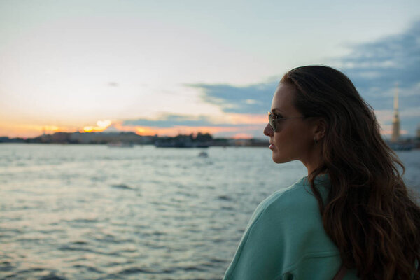 Young and beautiful girl sitting on the embankment of the river. she looks at the sunset and ships passing by. Saint Petersburg, Russia