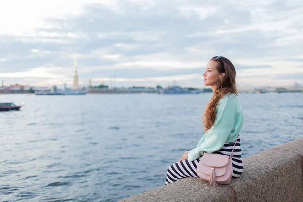 Menina jovem e bonita sentada no dique do rio. Ela olha para o pôr-do-sol e para os navios que passam. São Petersburgo, Rússia — Fotografia de Stock