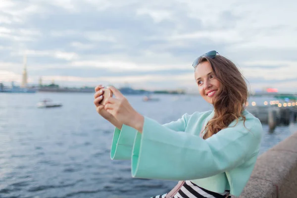Genç ve güzel kız nehir dolgu üzerinde oturuyor. o gün batımı ve bir selfie üstünde senin telefon alarak bakar. Saint Petersburg, Rusya Federasyonu — Stok fotoğraf